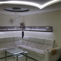 Nowoczesny salon z białym narożnikiem, szklanym stolikiem kawowym i podświetlanym sufitem podwieszanym z kryształowym żyrandolem.