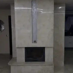 Obudowa kominka wyłożona beżowymi płytami imitującymi trawertyn, z pionowym pasem dekoracyjnym z mozaiki szklanej, widok na salon.