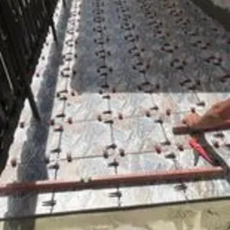 Układanie płytek na balkonie z użyciem systemu poziomowania, widoczne krzyżyki dystansowe i poziomica, w tle balustrada i zieleń.