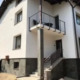 Dwupiętrowy dom z nową, białą elewacją i ciemnoszarą podmurówką. Balkon z czarną balustradą na pierwszym piętrze. Schody wejściowe z metalową barierką po prawej stronie.