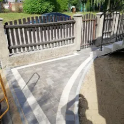 Świeżo ułożona, szara nawierzchnia z kostki, obramowana białymi elementami, prowadząca do drewnianej furtki w ogrodzeniu z betonowymi słupkami. Widoczny cień osoby fotografującej.