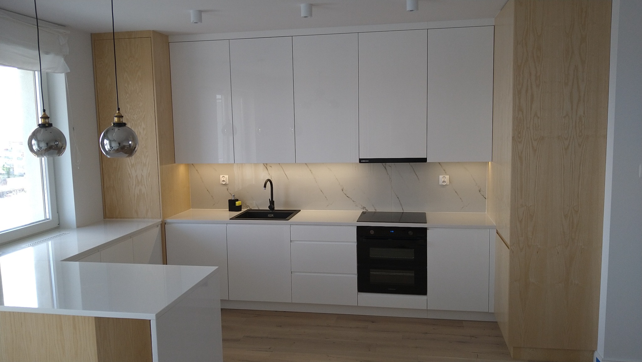 Nowoczesna kuchnia z białymi, lakierowanymi frontami szafek i elementami z jasnego drewna, czarny zlew i bateria, podświetlenie LED nad blatem z marmurowym wzorem, wiszące lampy w stylu glamour.