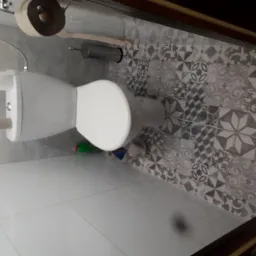 Nowoczesna toaleta podwieszana z szarą, wzorzystą okładziną ścienną i białymi płytkami podłogowymi, widoczny pojemnik na papier toaletowy i szczotka do WC.