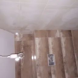 Świeżo położone, beżowe płytki na ścianie i suficie łazienki, widoczne instalacje wodne i oświetlenie montażowe.