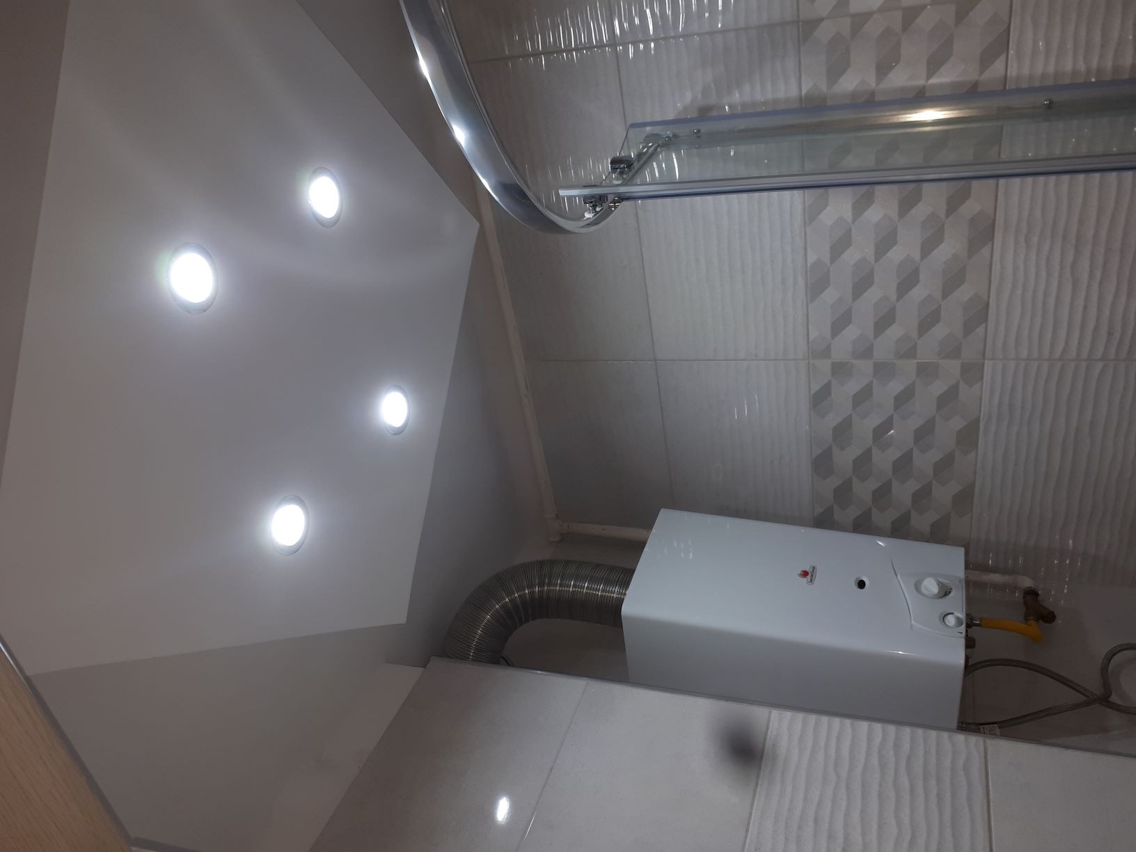 Nowoczesna łazienka z białymi płytkami, dekoracyjnym wzorem na ścianie, oświetleniem punktowym w suficie podwieszanym i zamontowanym piecykiem gazowym z rurą odprowadzającą spaliny.