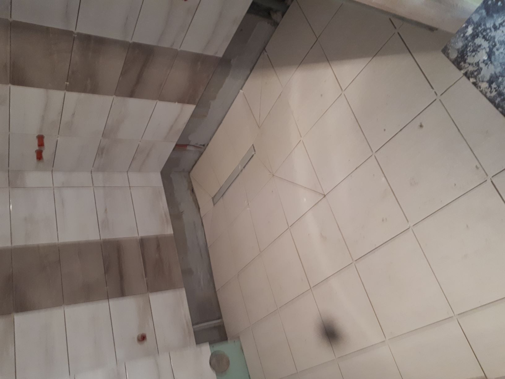 Ułożone płytki podłogowe i ścienne w łazience, widoczny odpływ liniowy w strefie prysznica, częściowo ułożone płytki.