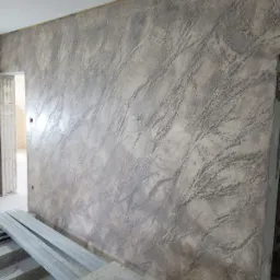 Ściana wewnętrzna z dekoracyjną, szarą strukturą przypominającą marmur, widoczne profile metalowe na podłodze, surowy stan wykończenia.