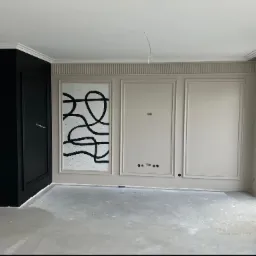 Wnętrze w trakcie prac wykończeniowych: ściany z dekoracyjnymi panelami i listwami, czarna zabudowa z drzwiami, surowa podłoga przed położeniem wykończenia.