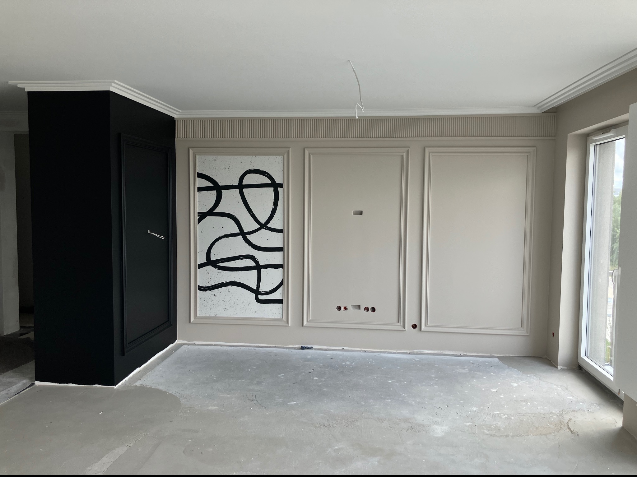 Wnętrze w trakcie prac wykończeniowych: ściany z dekoracyjnymi panelami i listwami, czarna zabudowa z drzwiami, surowa podłoga przed położeniem wykończenia.