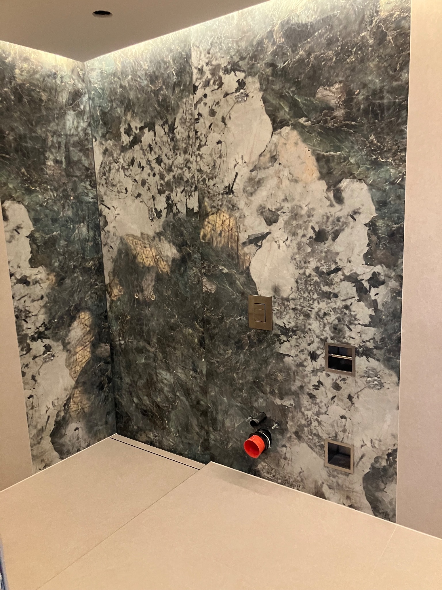 Wykończona łazienka z płytkami imitującymi marmur. Widoczne elementy instalacji wodnej i przycisk spłukiwania. Minimalistyczny design, beżowa podłoga.