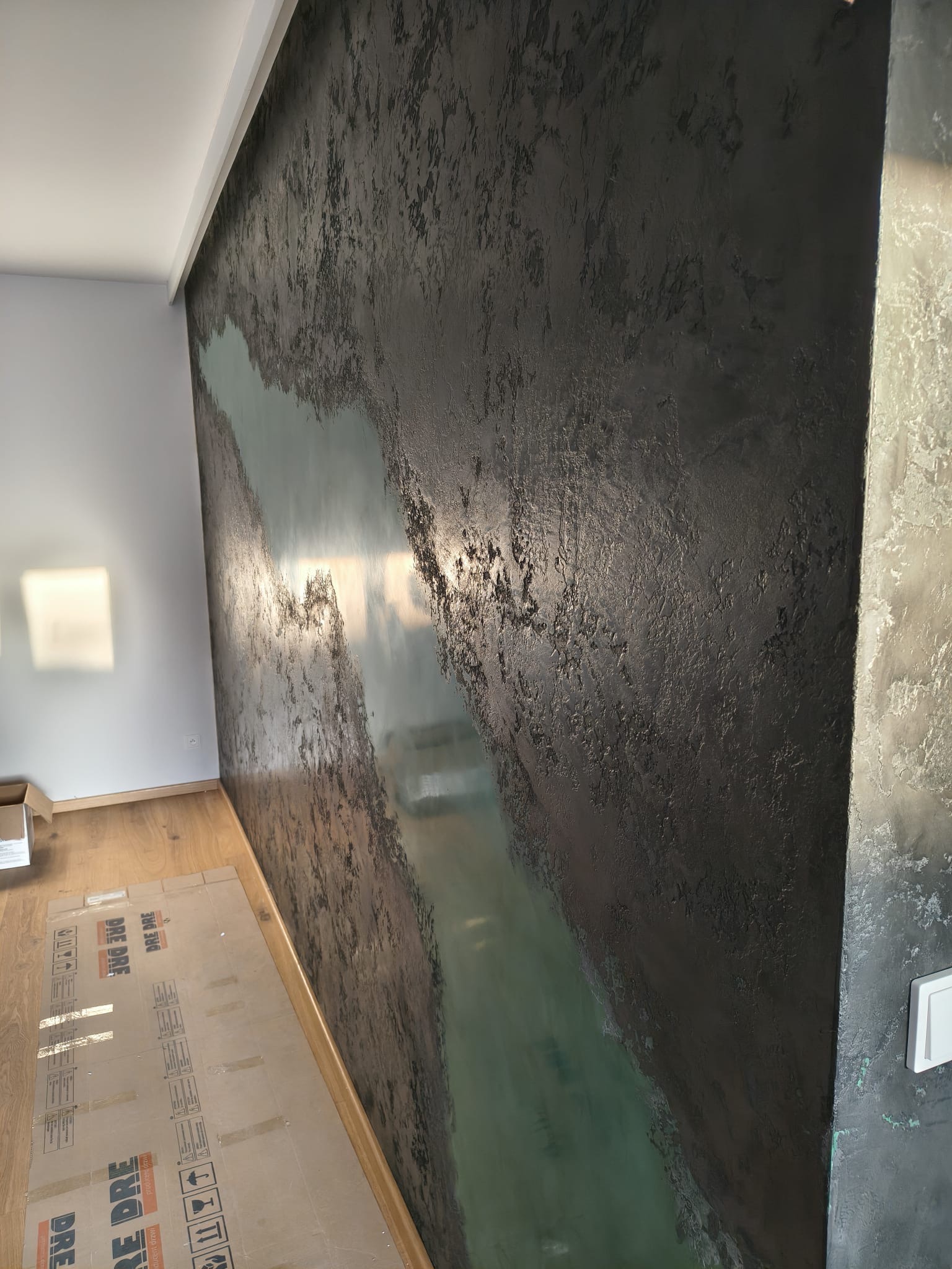 Dekoracyjna ściana z efektem rzeki w odcieniach szarości i zieleni, wykonana techniką strukturalną. Widoczne detale faktury i przejścia kolorystyczne.