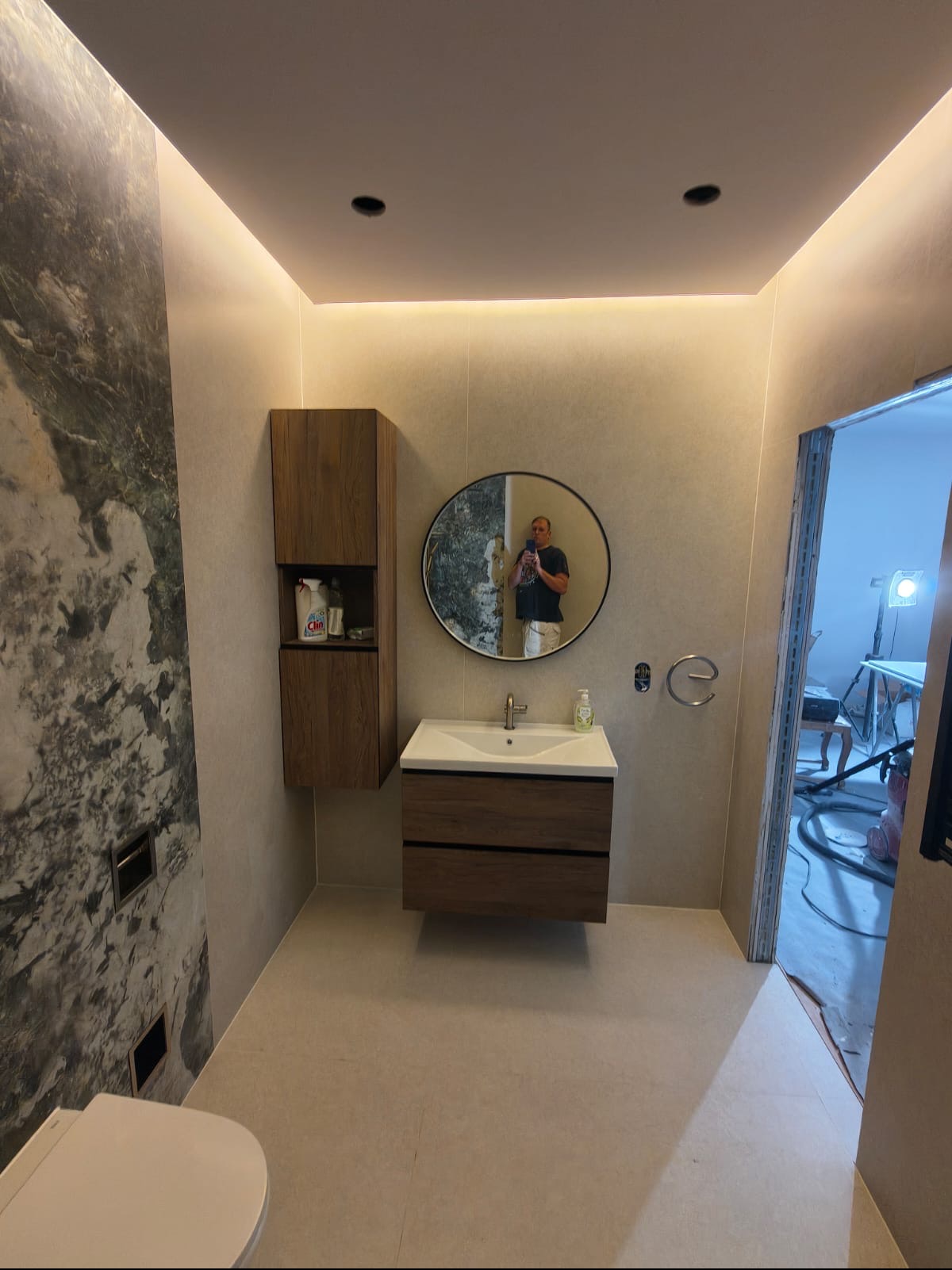 Nowoczesna łazienka po remoncie: umywalka z szafką, lustro w okrągłej ramie, marmurowa ściana, oświetlenie LED. W lustrze odbija się osoba robiąca zdjęcie.