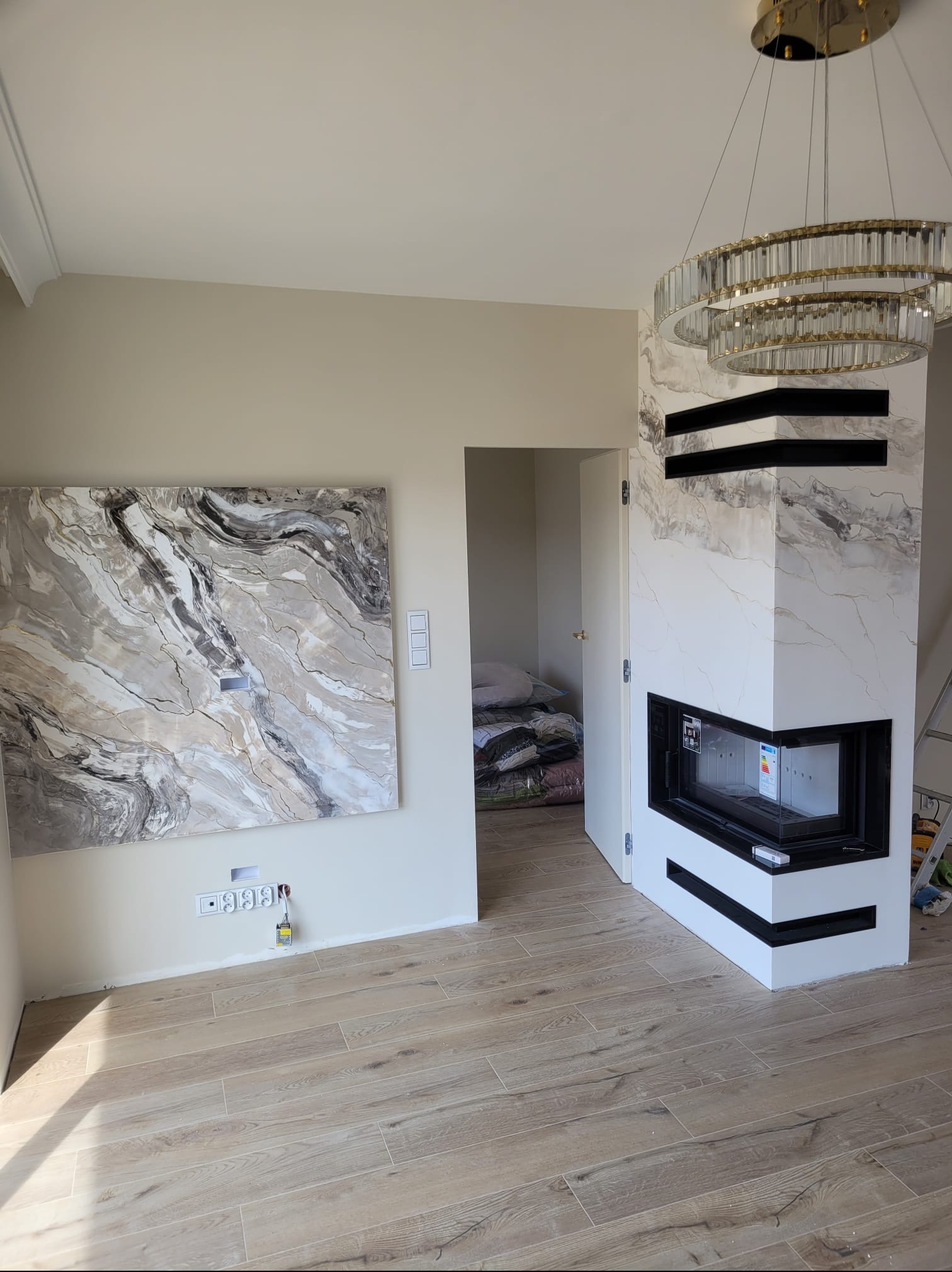 Salon po wykończeniu: marmurowy kominek z czarnym wkładem, abstrakcyjny obraz na ścianie, drewnopodobna podłoga i kryształowy żyrandol. Widok na otwarte drzwi.