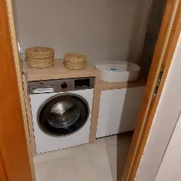 Pralka zabudowana pod blatem z dwoma koszykami dekoracyjnymi, obok umywalka nablatowa w szafce z szufladami, widok zza otwartych drzwi.