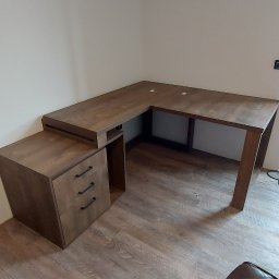 KATIE&JACK JACEK GACH - Brązowe biurko narożne z szufladami i otwartą półką, ustawione w rogu pokoju z panelami podłogowymi, widoczny fragment skórzanego fotela.