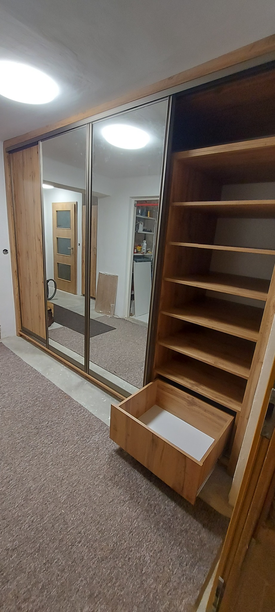 Drewniana szafa z przesuwnymi lustrzanymi drzwiami i otwartą półką z wysuniętą szufladą, widok na wnętrze pokoju z drzwiami i regałem.