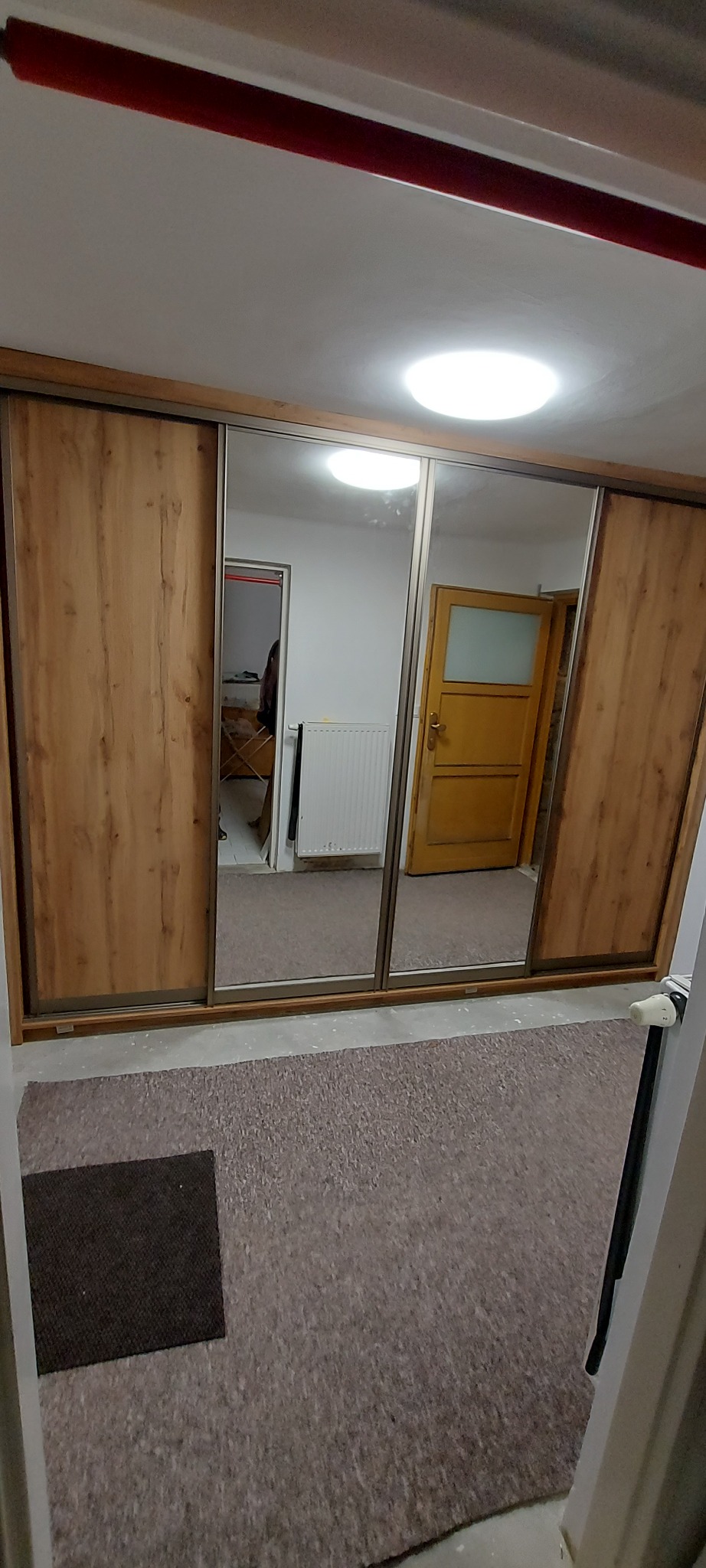 Trzydrzwiowa szafa przesuwna z lustrami, odbijająca wnętrze pokoju z grzejnikiem i otwartymi drzwiami.