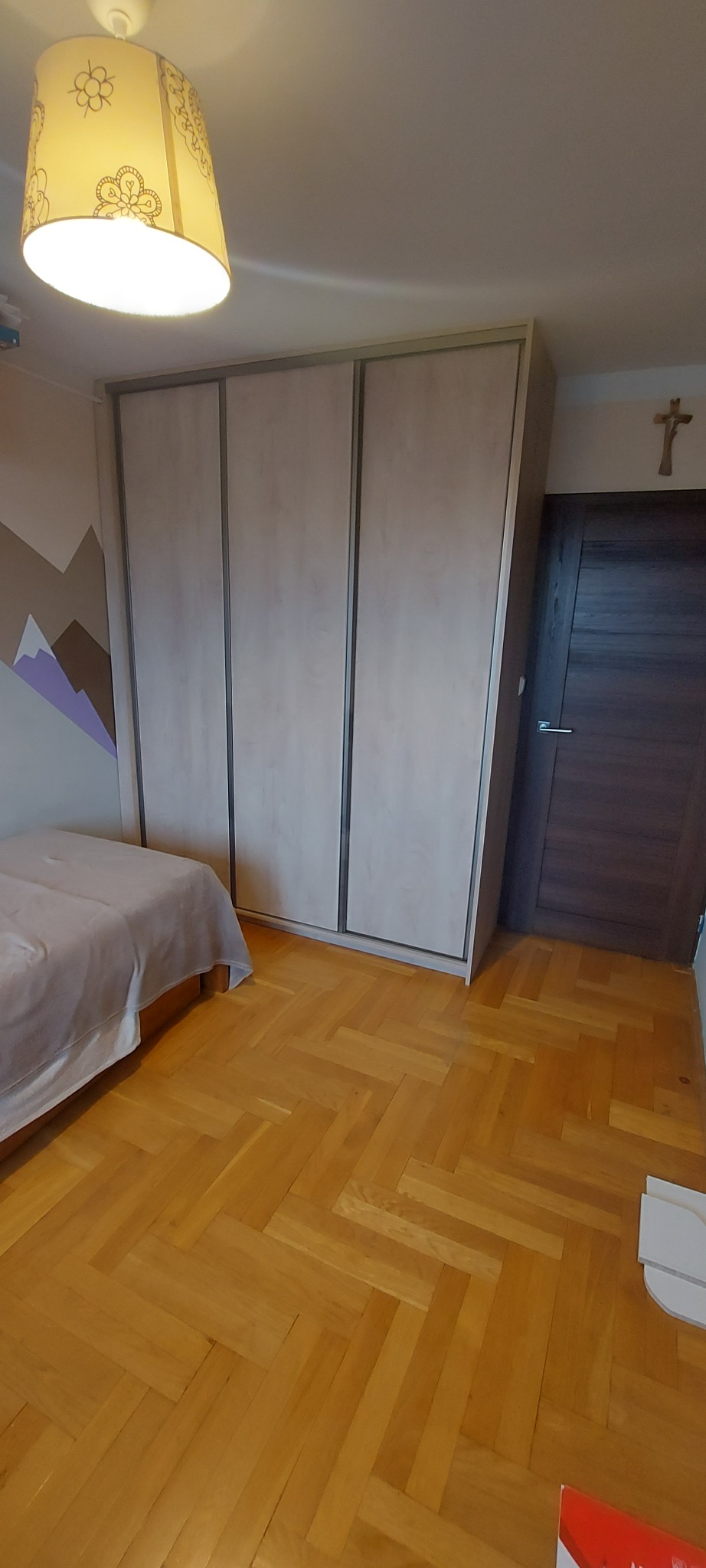 Jasna szafa przesuwna z trzema drzwiami, zamontowana w pokoju z drewnianą podłogą w jodełkę i łóżkiem z szarą narzutą, widoczny fragment ściany z malunkiem gór.