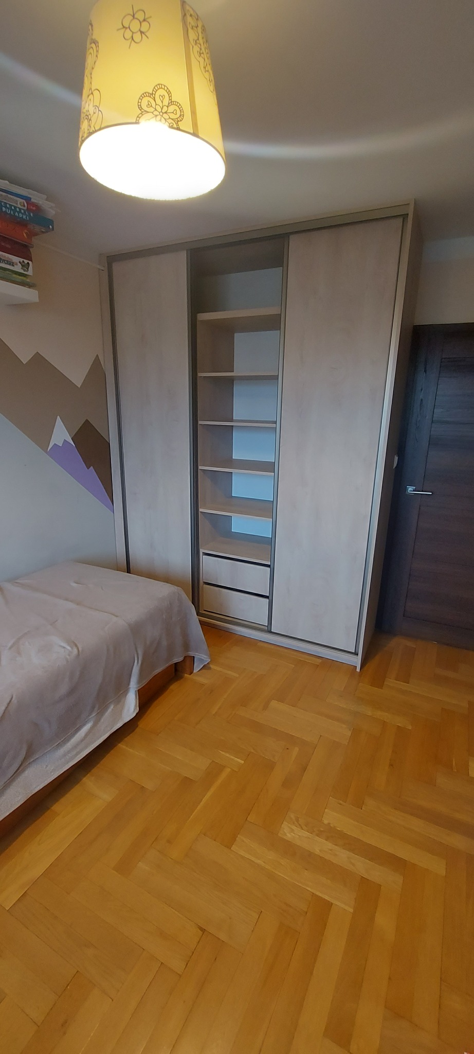 Jasna szafa przesuwna z otwartą częścią z półkami i szufladami, widoczna w pokoju dziecięcym z łóżkiem i malowanymi górami na ścianie.