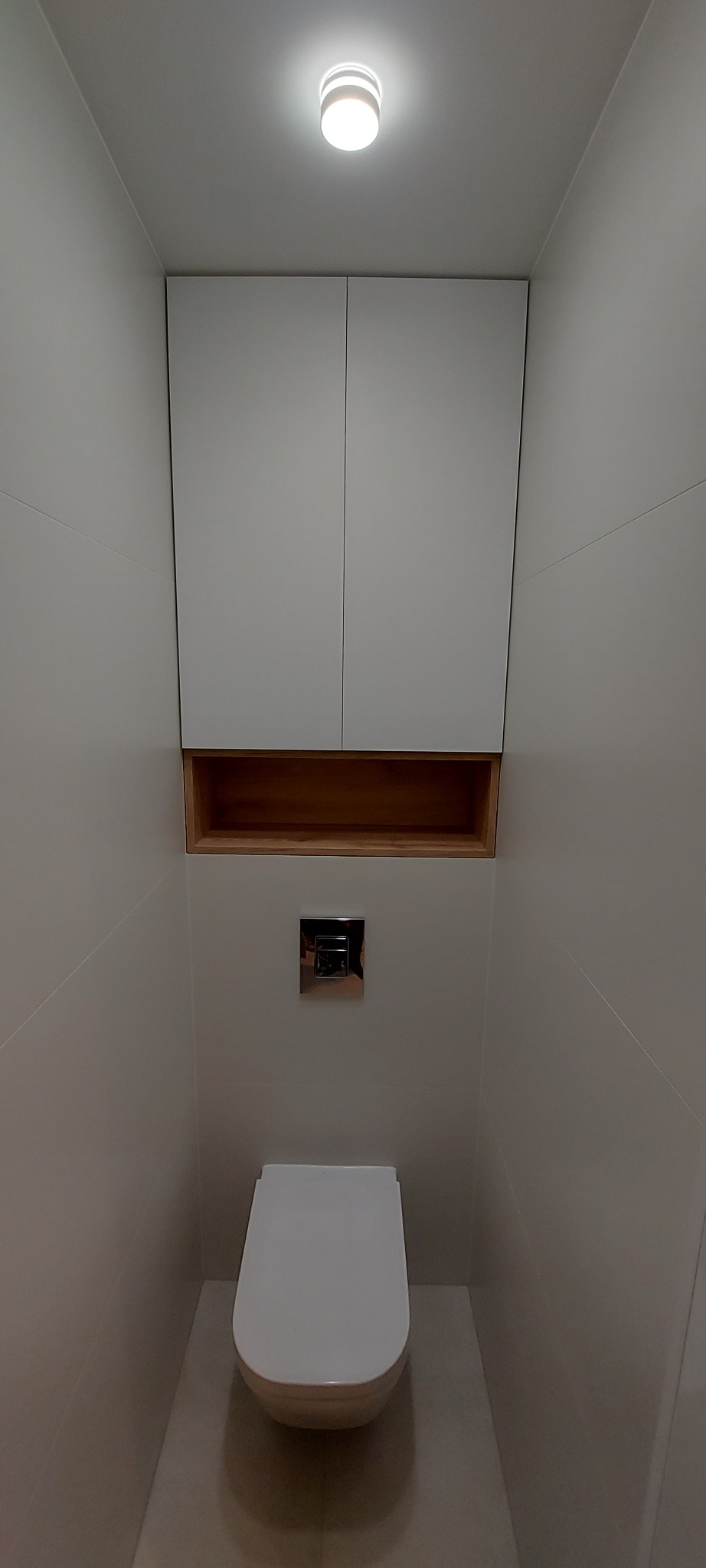Nowoczesna, wąska łazienka z podwieszaną toaletą, szafką na wymiar nad nią i drewnianą półką, oświetlona punktowym światłem.