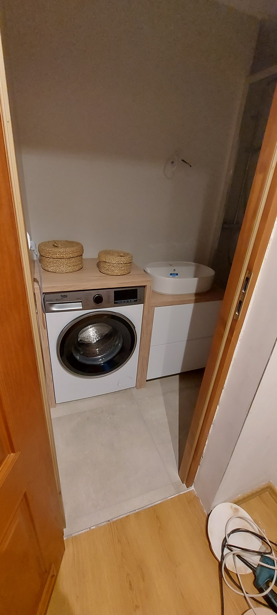 Pralka zabudowana pod blatem z dwoma koszykami dekoracyjnymi, obok umywalka nablatowa w szafce z szufladami, widok zza otwartych drzwi.