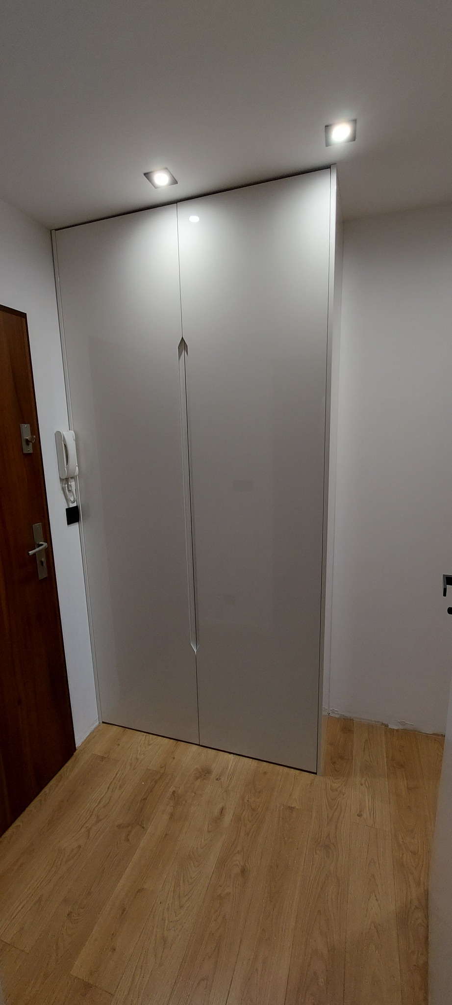 Biała, wysoka szafa z dwiema parami drzwi z minimalistycznymi uchwytami w przedpokoju, oświetlona dwoma punktowymi lampami sufitowymi, z widocznym fragmentem drewnianej podłogi.