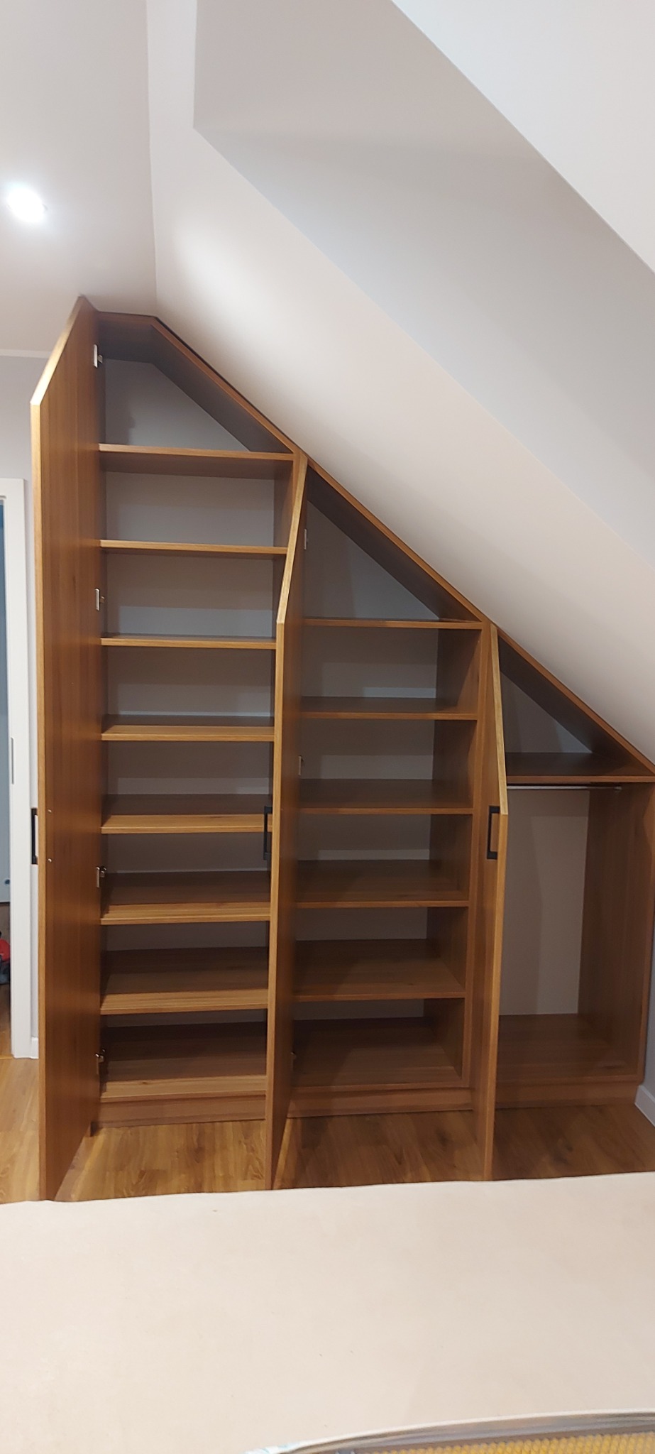 Drewniana szafa z półkami i drążkiem na ubrania, dopasowana do skosu dachu, z otwartymi drzwiami ukazującymi wnętrze.