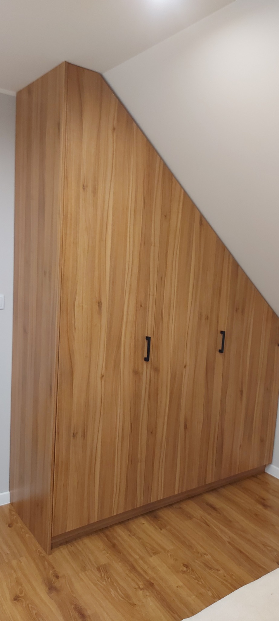 Trzydrzwiowa szafa z forniru o naturalnym wzorze drewna, dopasowana do skosu poddasza, z czarnymi, pionowymi uchwytami.