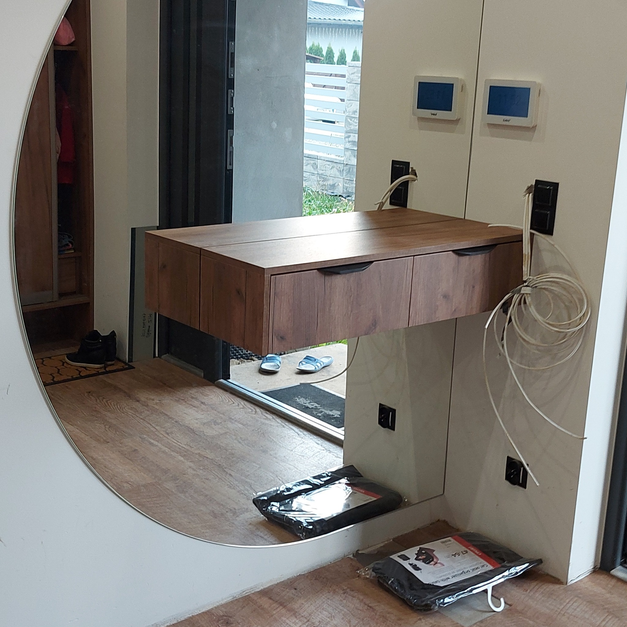 Nowoczesna, wisząca szafka z szufladami o drewnianym wykończeniu, zamontowana na białej ścianie z widocznymi kablami i regulatorami temperatury, odbicie wnętrza szafy i wyjścia na zewnątrz...