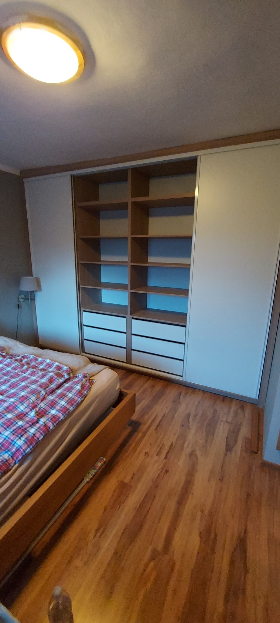 Jasna szafa z przesuwnymi drzwiami i otwartymi półkami w sypialni, widoczny fragment łóżka z czerwoną kratą, drewniana podłoga i sufit z okrągłą lampą.