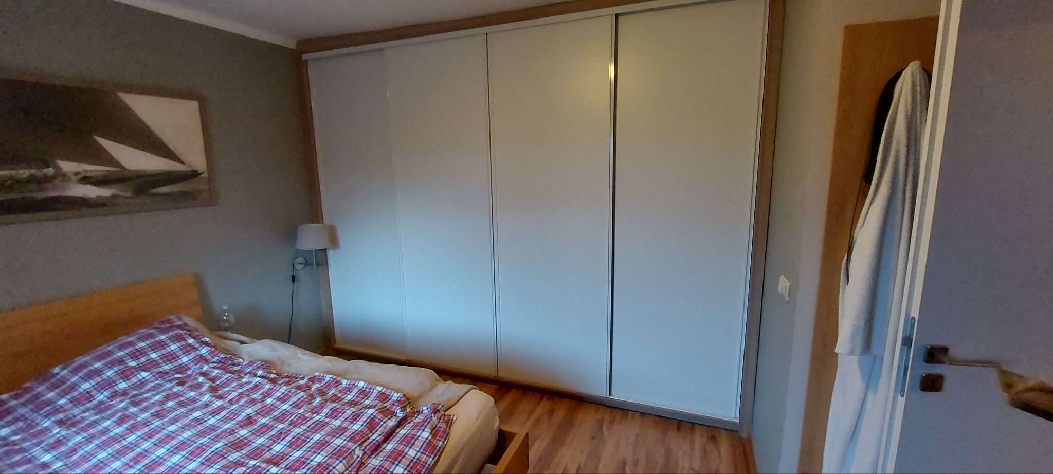 Biała szafa wnękowa z przesuwnymi drzwiami w sypialni, widok na łóżko z czerwoną kratą i obraz z motywem żaglówki na ścianie.