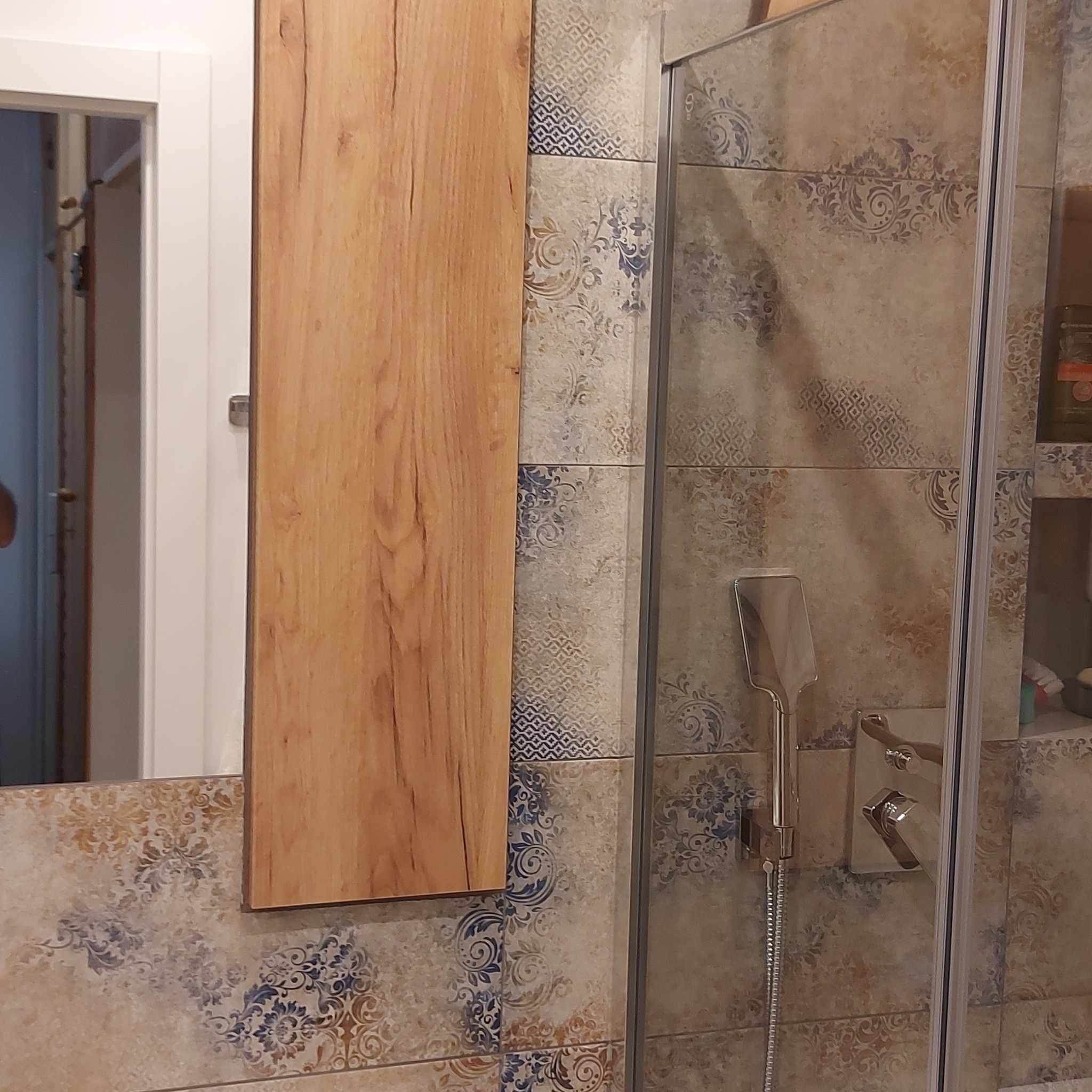 Drewniana szafka łazienkowa z lustrem, zawieszona obok kabiny prysznicowej z ozdobnymi, wzorzystymi płytkami.