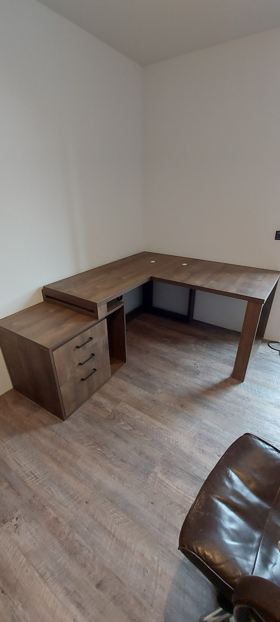 Brązowe biurko narożne z szufladami i otwartą półką, ustawione w rogu pokoju z panelami podłogowymi, widoczny fragment skórzanego fotela.