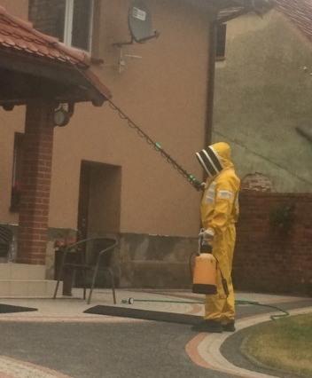 Osoba w żółtym kombinezonie ochronnym i masce pszczelarskiej spryskuje poddasze domu długą lancą z dyszą, trzymając zbiornik z preparatem w ręku.