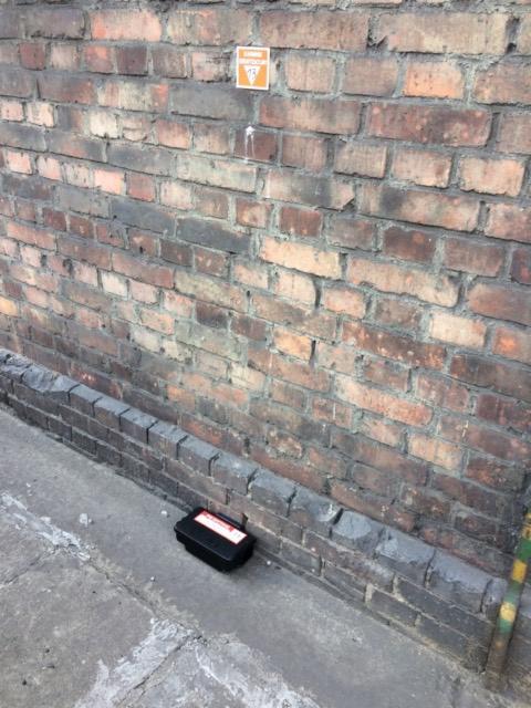 Czarna stacja deratyzacyjna ustawiona przy ceglanej ścianie z naklejką ostrzegawczą na górze, widok z góry.