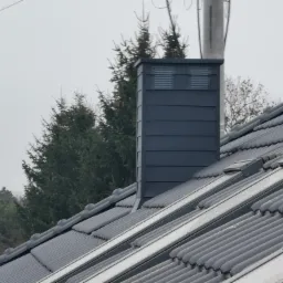 Szary dach z dachówki ceramicznej z czarnym kominem wentylacyjnym; widoczne okna dachowe i drzewa w tle.