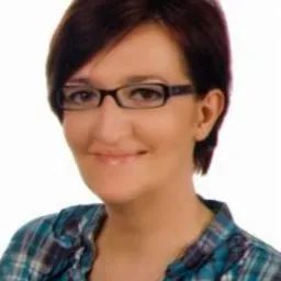 Portret kobiety w okularach z krótkimi brązowymi włosami, ubranej w niebiesko-zieloną koszulę w kratę i srebrny naszyjnik, uśmiechającej się delikatnie.