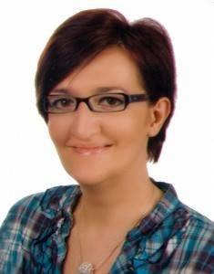 Portret kobiety w okularach z krótkimi brązowymi włosami, ubranej w niebiesko-zieloną koszulę w kratę i srebrny naszyjnik, uśmiechającej się delikatnie.