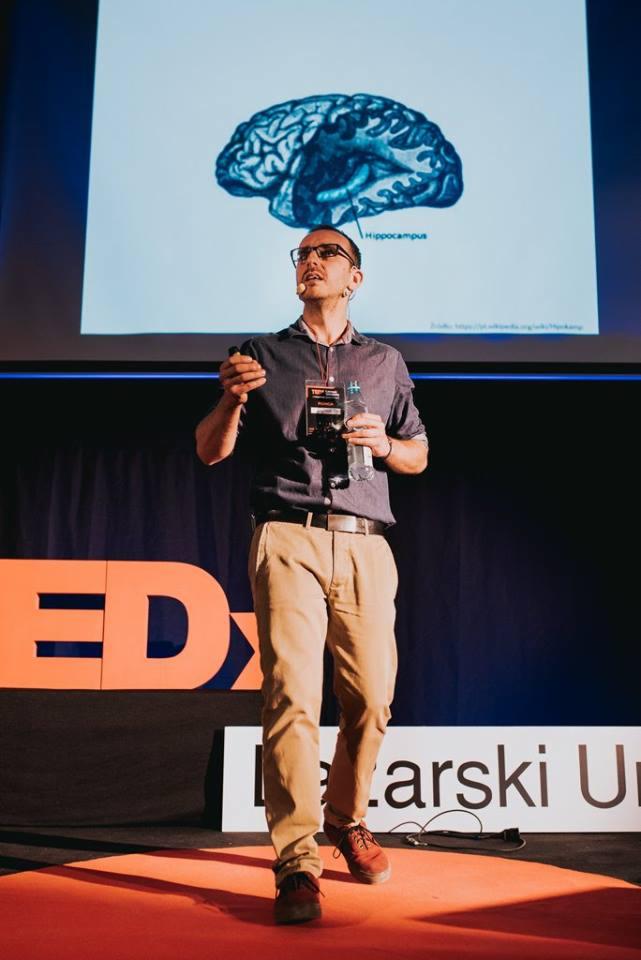 Prelegent w okularach prezentuje ilustrację mózgu z zaznaczonym hipokampem podczas konferencji TEDx.