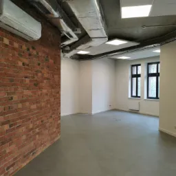 Surowe wnętrze biurowe z odsłoniętą czerwoną cegłą, szarą betonową podłogą, instalacją wentylacyjną pod sufitem i klimatyzatorem w folii ochronnej, świeżo po remoncie.