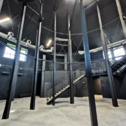 Wnętrze budynku z metalową konstrukcją nośną pomalowaną na czarno, widoczne schody metalowe, okna i instalacja wentylacyjna pod sufitem.