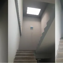 Klatka schodowa w szarościach z widocznymi grzejnikami, oświetleniem sufitowym i schodami wyłożonymi płytkami. W tle widoczna instalacja elektryczna.