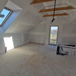 Poddasze w trakcie wykańczania: surowe drewniane belki stropowe, okno dachowe, ściany częściowo pokryte gładzią, na podłodze płyta OSB, widoczne instalacje i kartony z wyposażeniem łazienki.