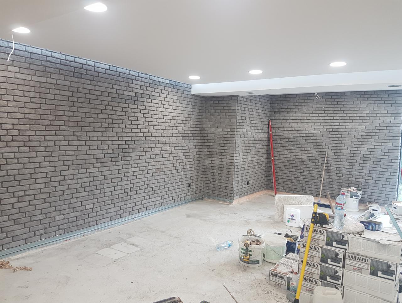 Pomieszczenie w trakcie prac wykończeniowych, ściany wyłożone dekoracyjną szarą cegłą, widoczne narzędzia i materiały budowlane, w tym pudełka z napisem HARVARD.