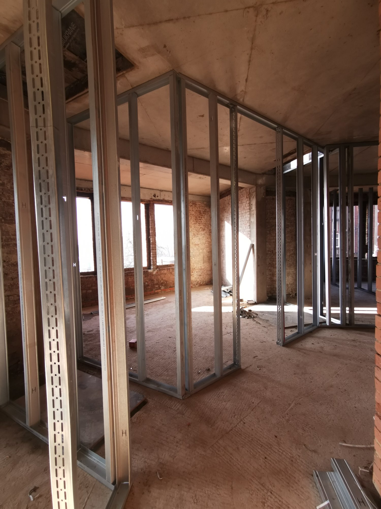 Metalowa konstrukcja ścian działowych z profili stalowych w surowym stanie, widoczne otwory w profilach, ceglana ściana w tle, przygotowanie do montażu płyt kartonowo-gipsowych.