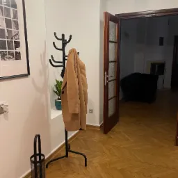 Wnętrze mieszkania z parkietem w jodełkę, wieszakiem z płaszczem, czarną ramą z grafiką na ścianie oraz otwartymi drzwiami do pokoju z kominkiem i czarnym fotelem.