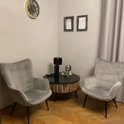 Wnętrze biura z dwoma szarymi fotelami, okrągłym stolikiem z napojami i lampą, dyplomami na ścianie i zegarem.