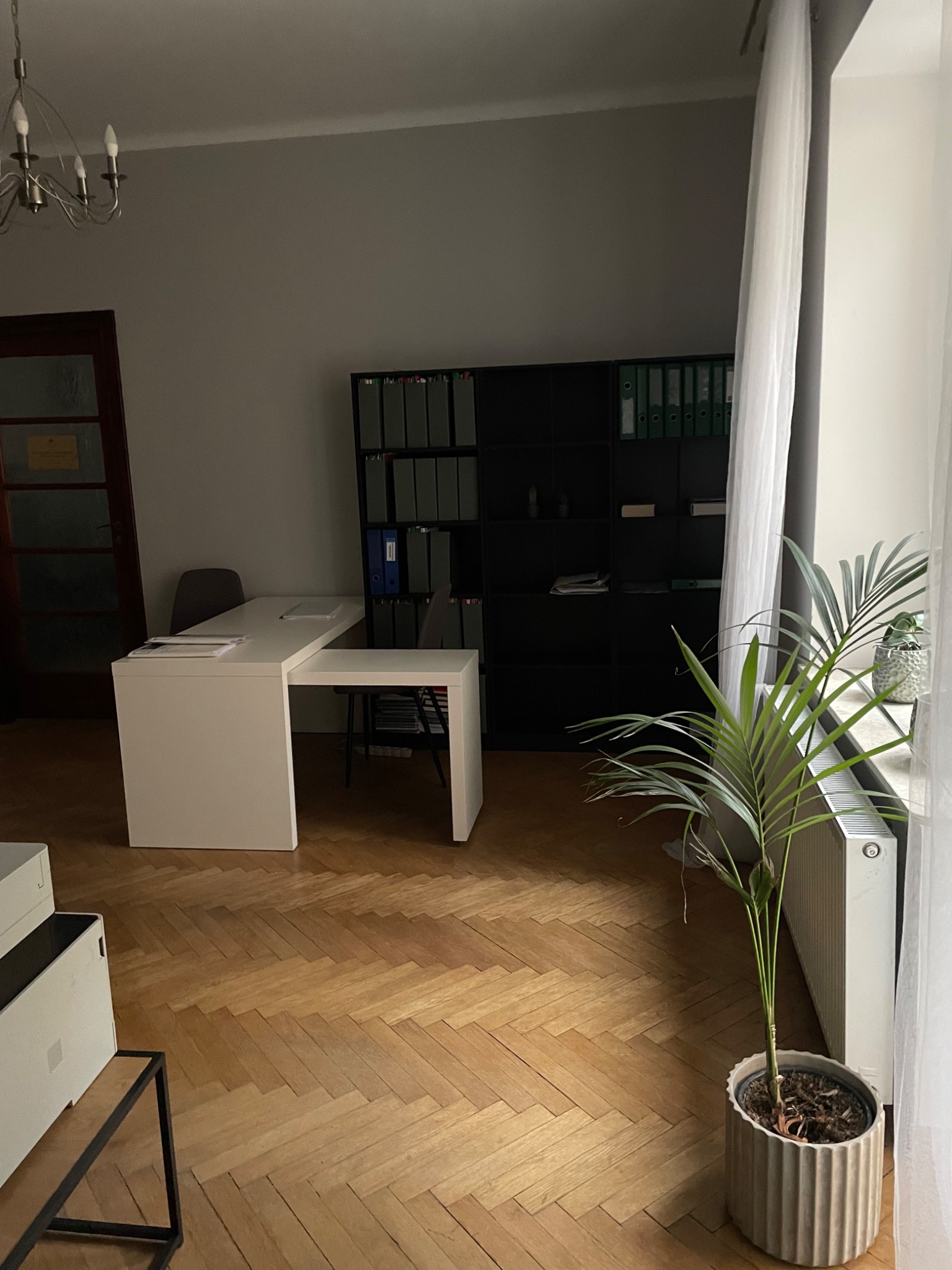 Wnętrze biura w Lublinie z białym biurkiem, regałem na dokumenty, parkietem w jodełkę i rośliną doniczkową przy oknie z białymi zasłonami.