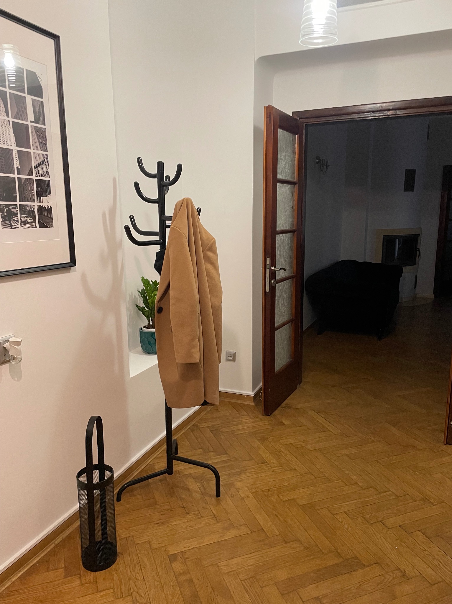 Wnętrze mieszkania z parkietem w jodełkę, wieszakiem z płaszczem, czarną ramą z grafiką na ścianie oraz otwartymi drzwiami do pokoju z kominkiem i czarnym fotelem.