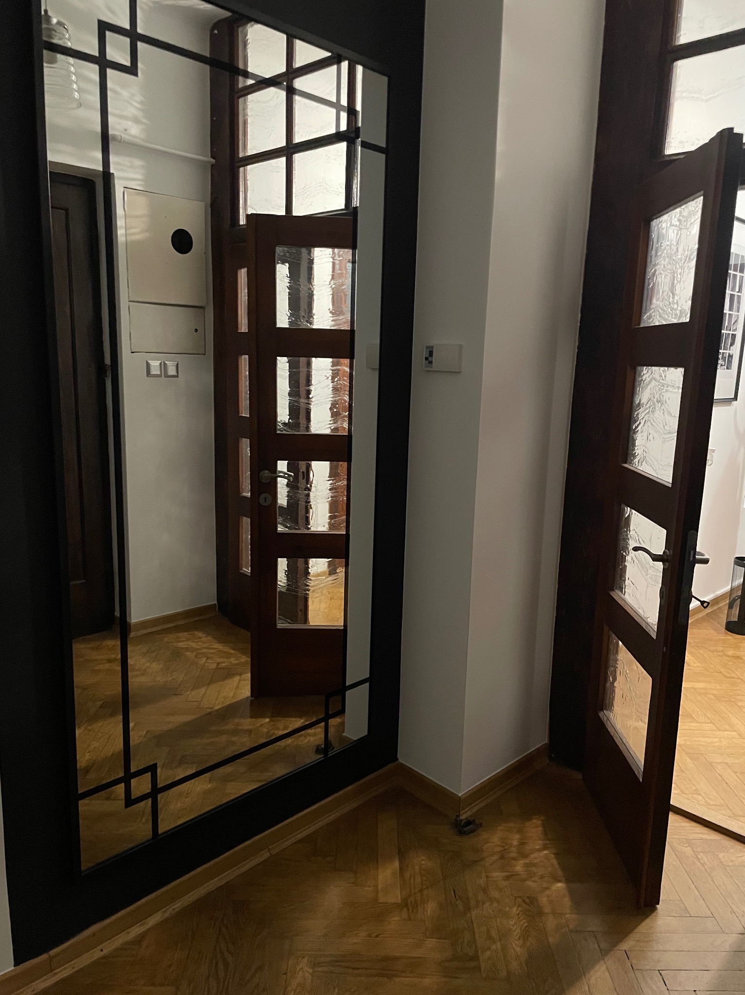 Wnętrze mieszkania z drewnianą podłogą w jodełkę, widoczne drzwi z szybami i duże lustro z czarną, geometryczną ramą.